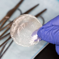 A gloved hand holds a quasi-transparent circular film over a tray of metal instruments