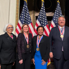 Several people in suits, wearing medals, standing in front of 3 U.S. flags