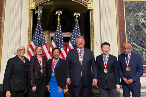 Several people in suits, wearing medals, standing in front of 3 U.S. flags